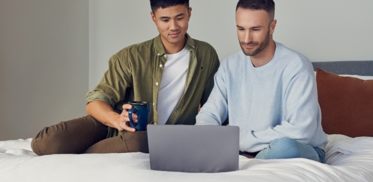 Zwei Männer sitzen entspannt auf einem Bett und schauen auf einen Laptop.